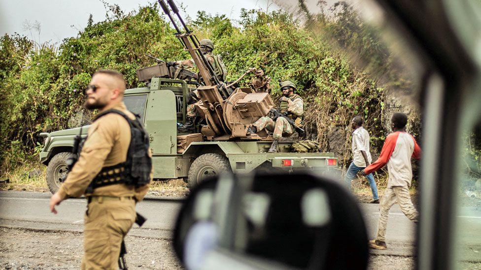 A photo taken from a vehicle showing an armed mercenary in military fatigues and a black backpack walking along a road north-west of Goma. Behind him is a Congolese army truck with a mounted gun. Two male civilians are also pictured walking by - 23 January 2025