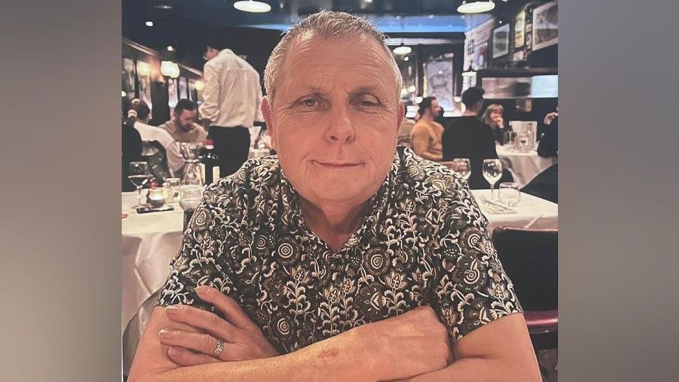 Dave Pickering sitting with his arms crossed. he is wearing a floral patterned black and white shirt, he is smiling