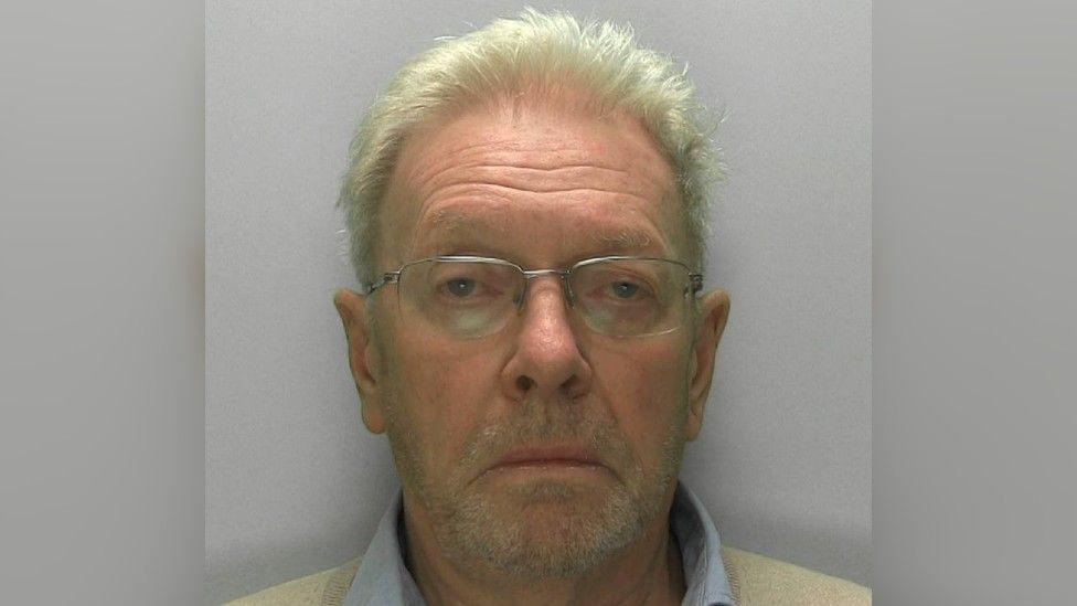 A police custody image of Dr Charles Ragan, taken against a grey background. He is pictured unsmiling, wearing thin-framed glasses, a pale collared shirt and a light coloured jumper.