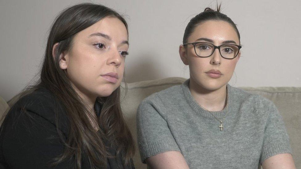 Cara and Elena Lang sit on a sofa together. Cara, who has long brown hair, looks to the side while her sister, Elena, whose dark hair is tied back, looks into the camera