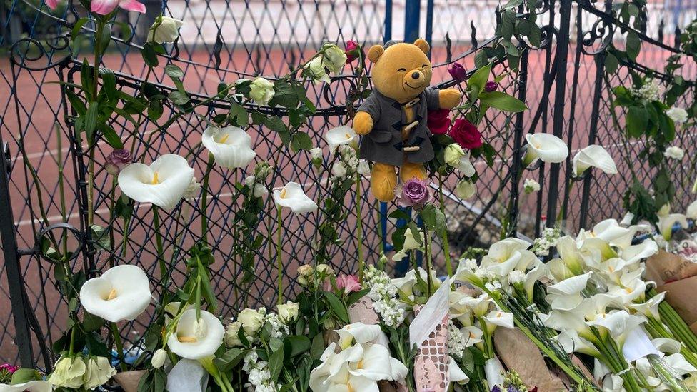 A bear hangs on a fence with white lilies below