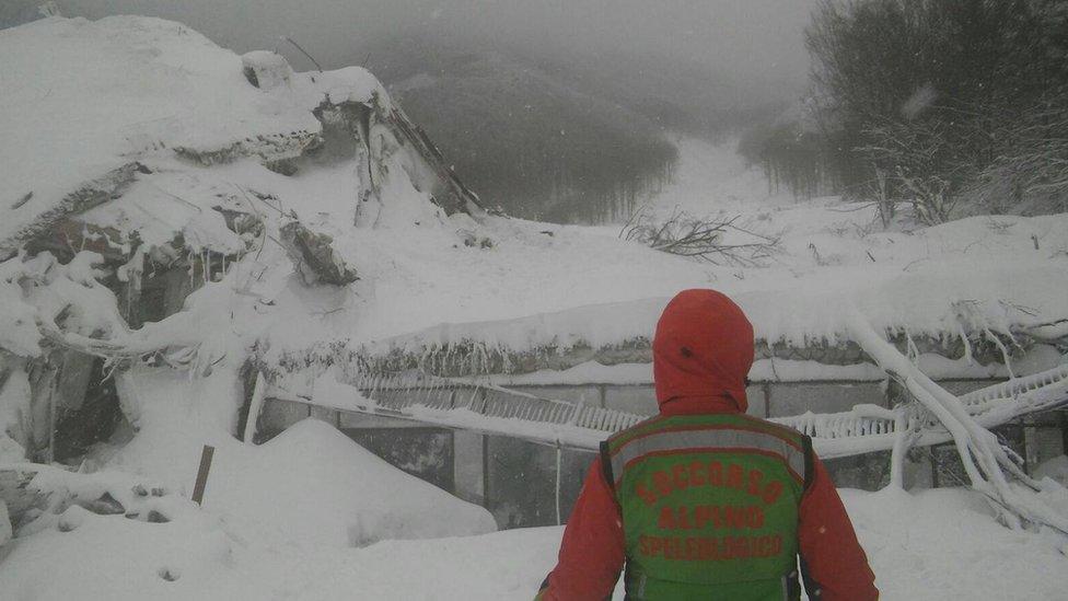 A man in high-visibility snow gear stands before a mound of white snow, under which a bare hint of a building can be seen.