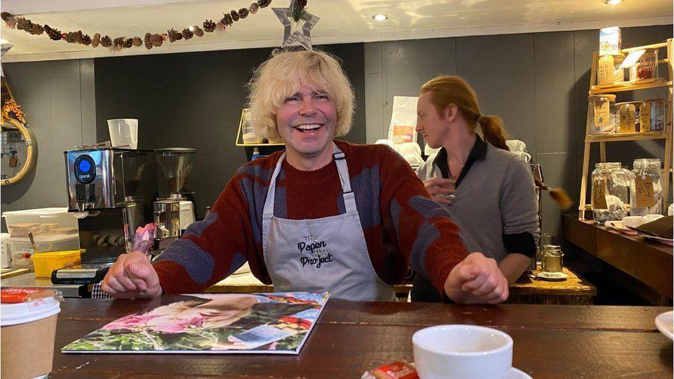 Tim Burgess, with long blonde hair and wearing a red and blue jumper, smiles behind the counter of a cafe while wearing a Paper Cup Project overall.