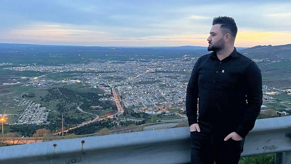 Kardo Jaf, i njohur edhe si Kardo Ranya, qëndron në një pikë vrojtimi buzë rrugës pranë një parmaku metalik mbrojtës, i veshur me një këmishë të zezë me mëngë të gjata dhe pantallona të errëta, me duart në xhepa dhe trupin e kthyer pak djathtas. Pika e vrojtimit shikon një peizazh të gjerë qyteti të luginës në muzg, me grumbuj ndërtesash, rrugë dredha-dredha dhe drita rrugësh të shpërndara që shtrihen përgjatë kodrave të gjelbra. Qielli është blu i zbehtë me re të buta dhe dritë dite që po venitet, dhe kodra të largëta formojnë horizontin në sfond.