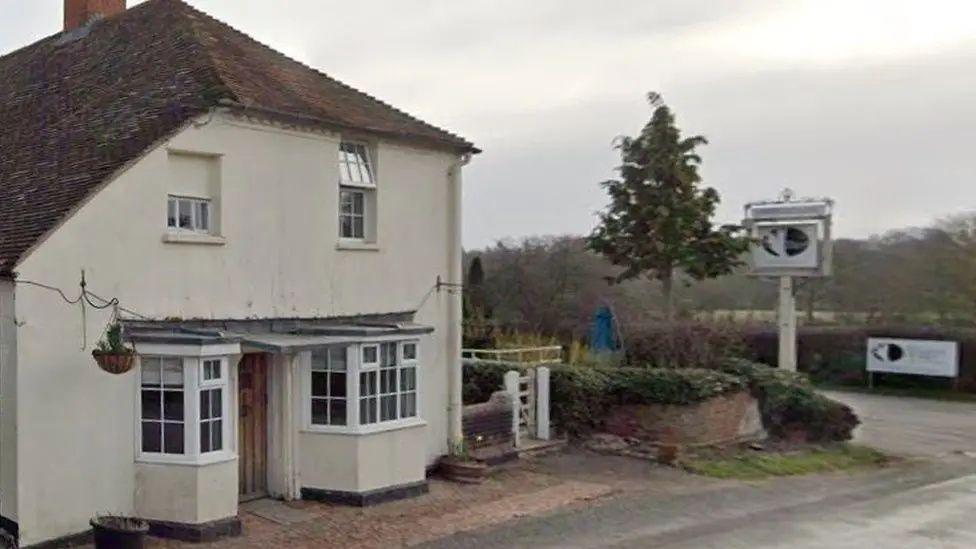 A google maps image of the outside of The White Hart Inn which has a white front, wooden door and unusually shaped roof which slants unevenly on one side