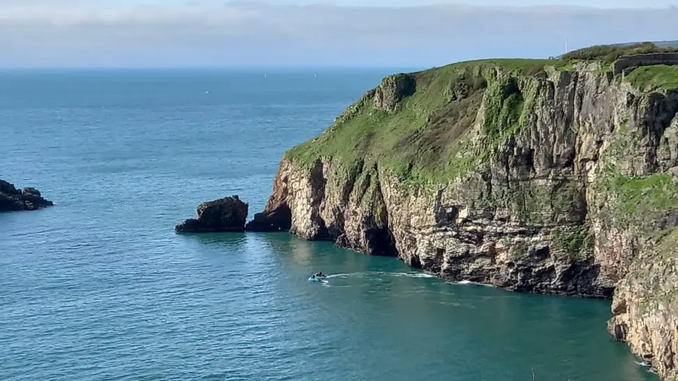 Berry Head is a grassy steep-sided headland with rocky cliffs surrounded by the sea. At its end is a large rock and a small distance away from it is an island. There are two jet skis off the rocky coastline. It is a clear sky day and the blue sea is calm.
