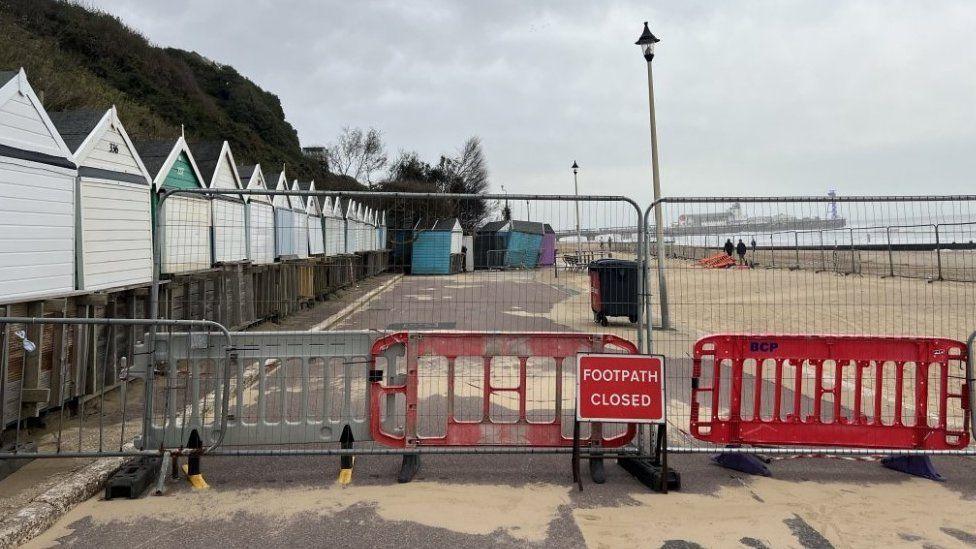 Metal and plastic fencing across path with beach huts to the left, sandy beach to right and pier in background