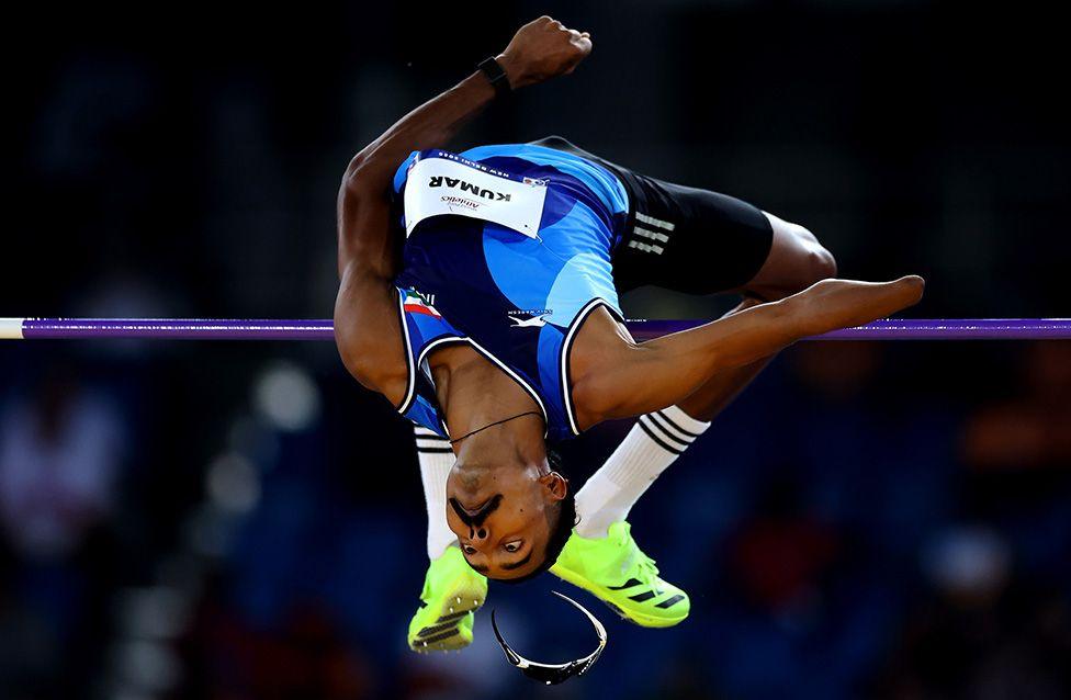 Nishad Kumar wins the gold medal in the Men’s High Jump T14 Final during day seven of the World Para Athletics Championships in New Delhi. The image shows him mid-air, arching backward over the bar in a blue and black uniform with bright neon green shoes, as stadium seating blurs in the background.