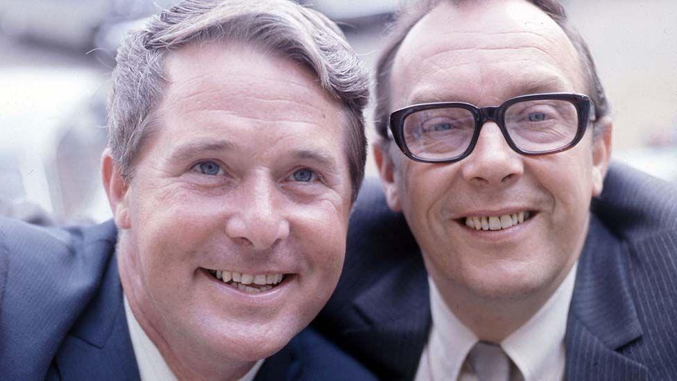 Close up image of smiling Ernie Wise and Eric Morecambe. Both are wearing blue suits