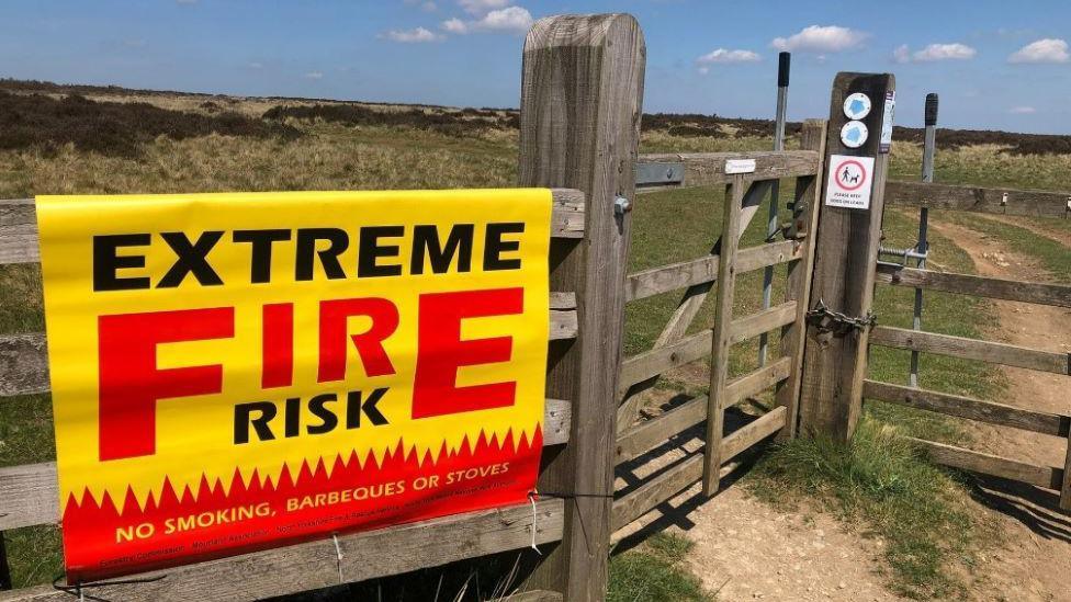 A yellow and red sign has been put up on a wooden fence and gate in a field, saying "Extreme fire risk. No smoking, barbecues or stoves". 