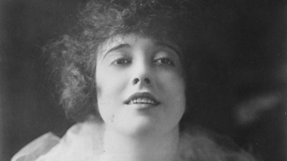 A black and white photo of Mabel as she stares directly into the camera with a small smile. Her hair is dark and curly and frames her face.