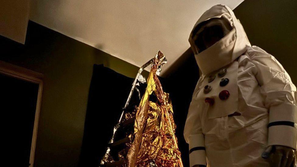 A person wearing a white spacesuit and space helmet. There is a ladder behind them, which is wrapped in gold-coloured material.