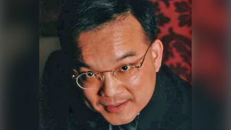 Kenneth Law has black hair and is wearing a black jacket. He is wearing glasses with thin rims and circular lenses.