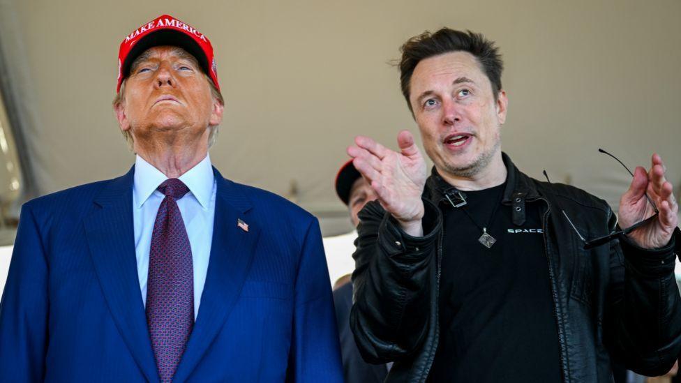 Donald Trump, an elderly man with orange face make-up, wearing a blue suit and red hat, stands next to Elon Musk, with dark hair and wearing all black. They are photographed from a low angle in a medium-close-up. 