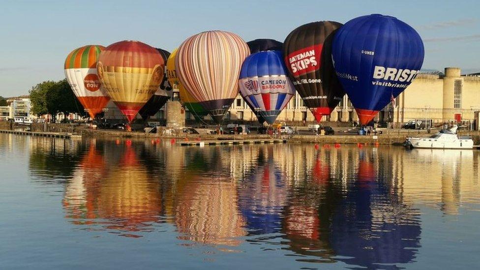 Nine colourful hot air balloons are inflated on the ground in front of the water