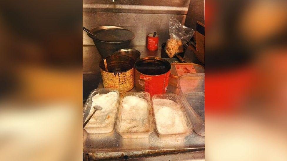 A silver kitchen surface covered with open, dirty containers - three containing what looks like salt and others containing sauce and oils.