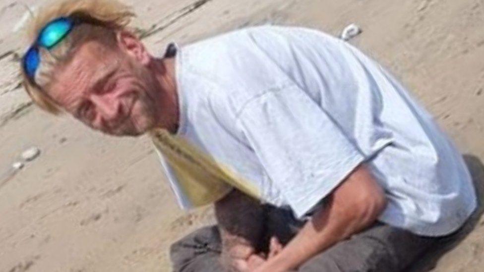 Man with blonde hair and sunglasses on his head looking at the camera he is wearing a grey t-shirt and dark trousers and sits cross-legged on a sandy beach.