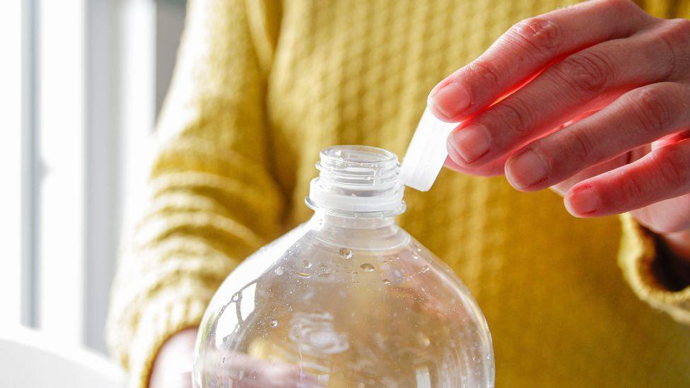 A person wearing a yellow jumper holding a clear plastic bottle.