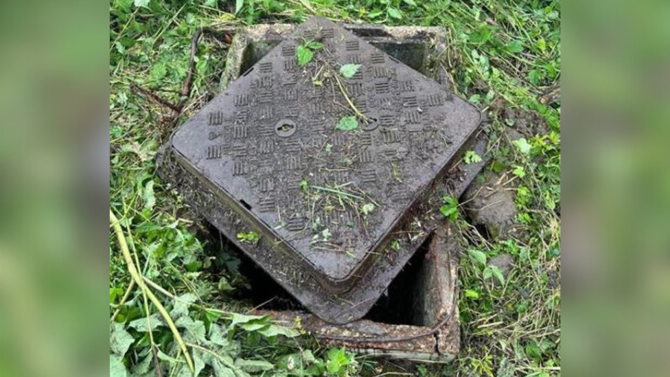 The manhole and its metal cover, which has been placed across the manhole rather than secured on. Grass surrounds the manhole.