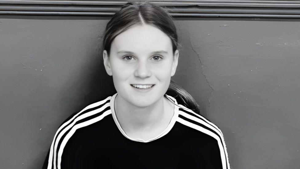 Black and white close-up of Holly Newton, smiling at the camera. Her long hair is tied back in a ponytail and she is wearing a dark t-shirt with lighter stripes on the arms and shoulder.