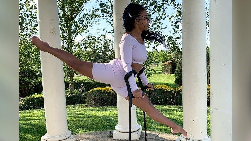 Sophia Adzoa-Moore looking away from the camera in a light pink matching long sleeve top and shorts set. She is dancing in the air and using a pair of crutches.