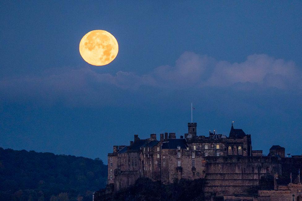 The full October moon, known as the Hunter's sets behind Edinburgh Castle, Scotland. 17 October  2024.   The moon is on the left hand side of a blue dusk sky. The castle in in the foreground to the right.