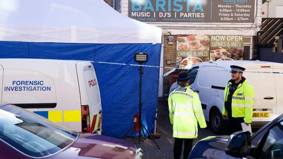 Police officers outside a building with a sign which reads BARISTA. There is a blue and white police tent, a police cordon and a police car which reads FORENSIC INVESTIGATION.