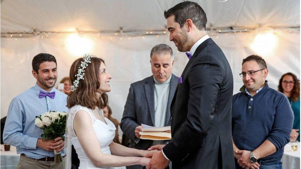 A man and woman, dressed in traditional wedding attire, hold hands and grin at each other as they get married.