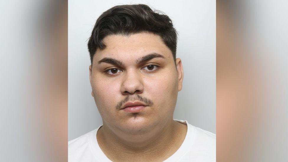 A police mugshot of Adi-Petronel Petrechiva, a young man with short black hair and a pencil moustache. He is wearing a white top.
