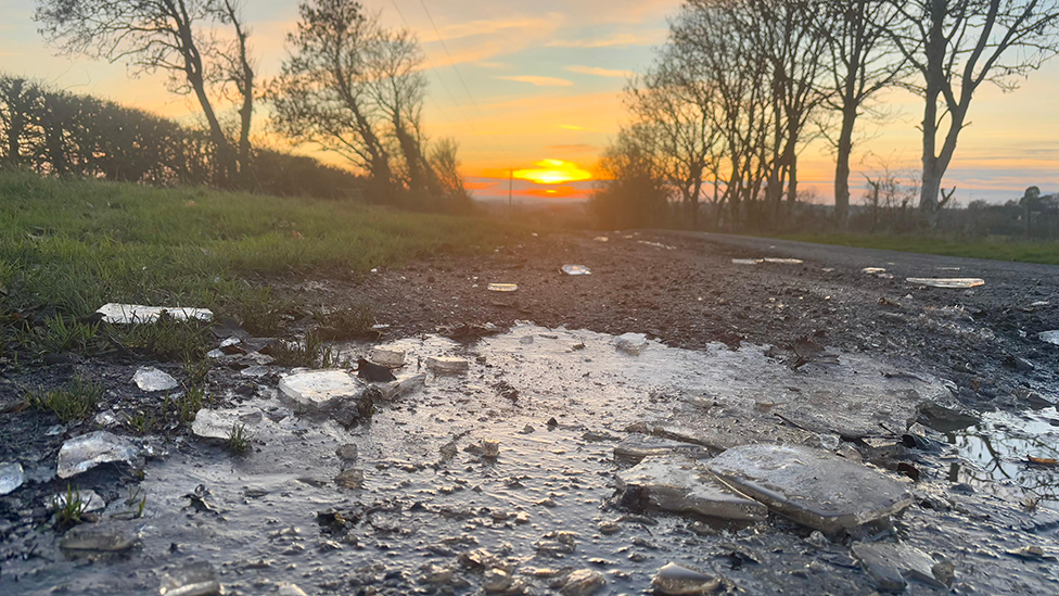 Sunset in Fenny Compton. Large chunks of ice lie on the side of a country road, mixing with the mud. A grass verge to the left is bordered by a hedge