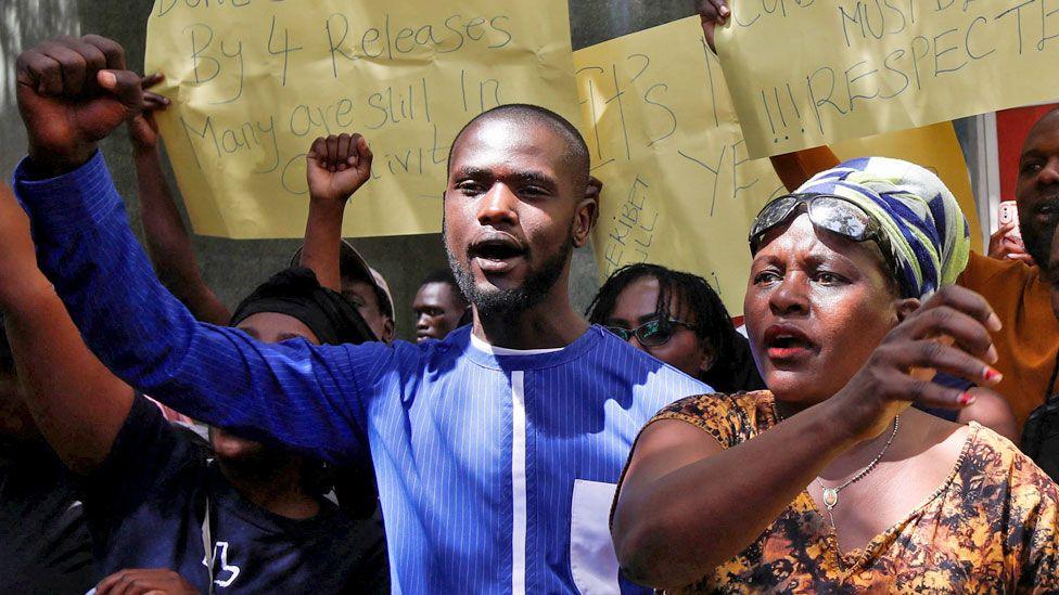 A of group of Kenyan protesters holding yellow hand-written posters aloft calling for the release of people who have been abducted.  Several - including a young bearded man in a blue collars shirt and a woman wearing a brown patterned V-neck dress, necklace, blue-and-white headscarf and sunglasses on her forehead in close-up - have their fists raised.  