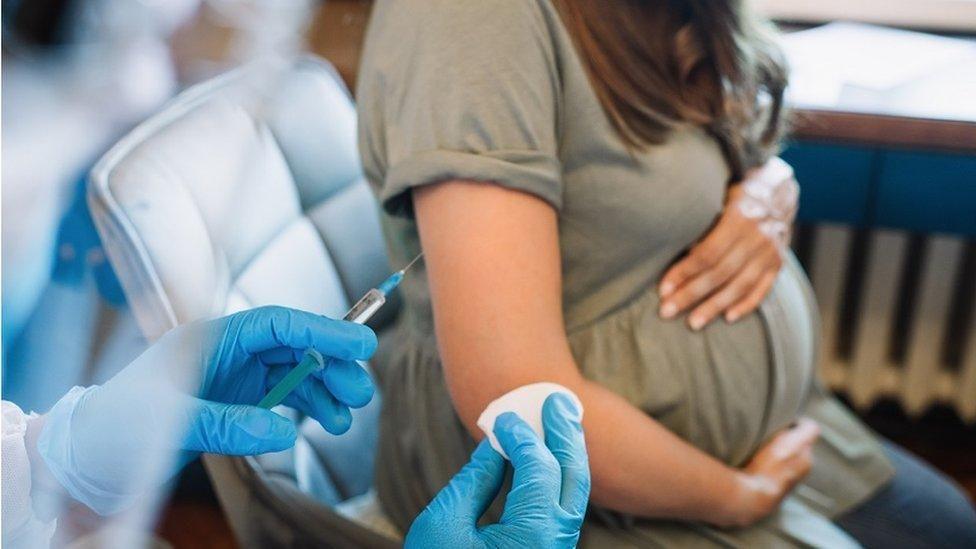 A pregnant woman in a green dress, holding her bump, sat in a grey chair, while the hands of a medical professional in blue gloves prepares a vaccine. 