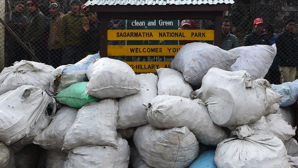 Locals look at piles of rubbish bags lying against an airport fence next to a sign reading "clean and green: Sagarmatha National Park Welcomes You"