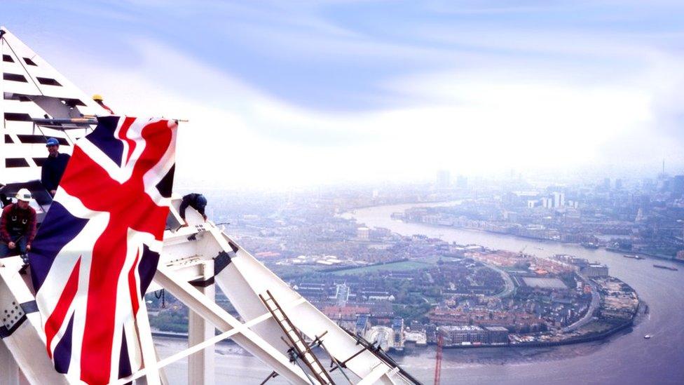 A photograph showing steel workers attaching the pyramid top to the skyscraper, with the River Thames and city behind.