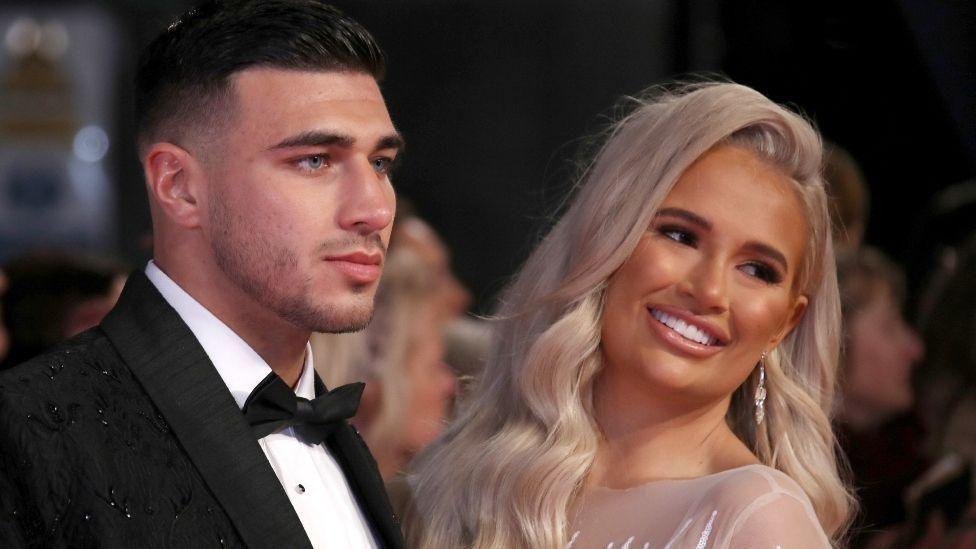 Tommy Fury, wearing a black tuxedo with white shirt and black bow-tie, and Molly-Mae Hague, wearing a sheer dress and with long blonde hair, looking off camera - Molly-Mae is smiling 