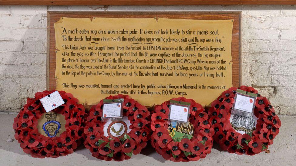 Four red poppy wreaths in a row resting underneath a wooden plaque against a wall to mark VJ Day in 2024. The plaque explains how a Union Flag was mounted, framed and created into a war memorial to remember men from the 4th Btn the Suffolk Regiment who died in World War Two in the far east. 