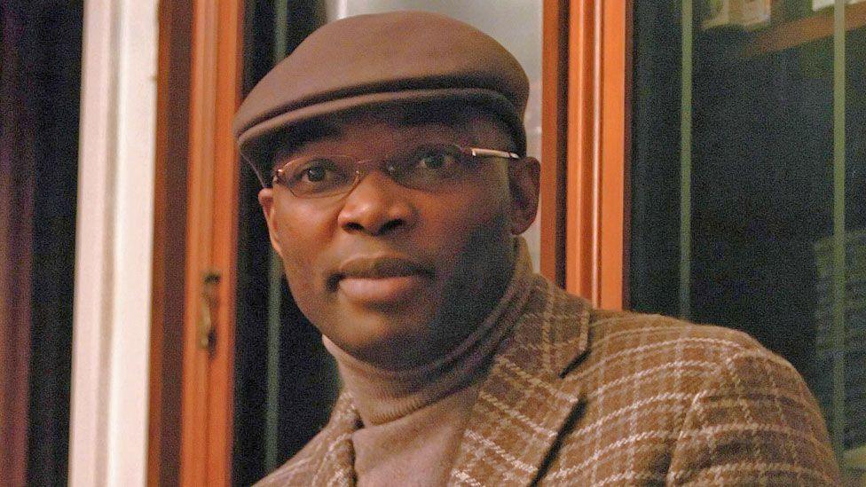 Charles Onana photographed in France in 2006 wearing a brown cap, a brown check jacket and beige turtleneck jumper