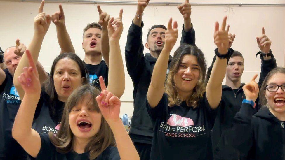 A close up of group of young men and women with black t-shirts holding their arms up in a dance hall. 