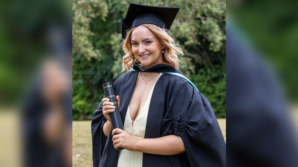 A portrait photo of a woman with shoulder length blonde hair which has moved back a bit in the wind. She is smiling and wearing a low light-yellow dress with a gold necklace and a black graduation gown, cap and rolled up certificate in a box.