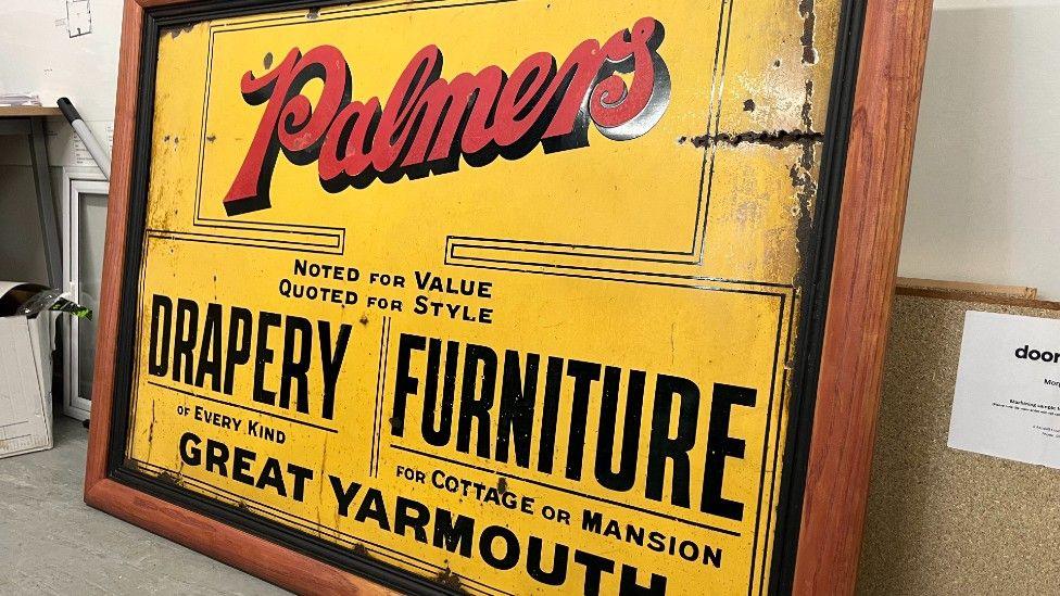 A yellow sign with a wooden frame. On the sign if red writing which says Palmers. There is also black writing on the sign which says Drapery and Furniture.