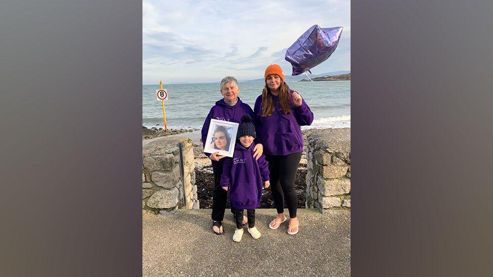 Sian Ashcroft’s mum, Sue Dowling, sister Lauren Dowling and Lauren’s son stand in front of a wall above the sea on Traeth Bychan beach in Anglesey. They wear purple jumpers; The mum holds a framed picture of her daughter. Lauren Dowling holds a purple star-shaped balloon.