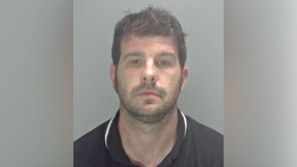 Stuart Worby police mugshot. He is a man with black hair and stubble wearing a black polo shirt