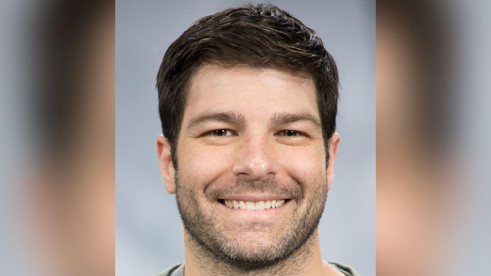 A man with short, neat black hair smiles widely in front of a grey background in a professional-looking headshot.