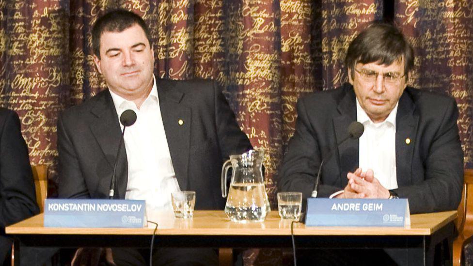 Konstantin Novoselov and Andre Geim sit a table with a jug of water and signs bearing their names. The physicists are sitting in front of a colourful curtain. 