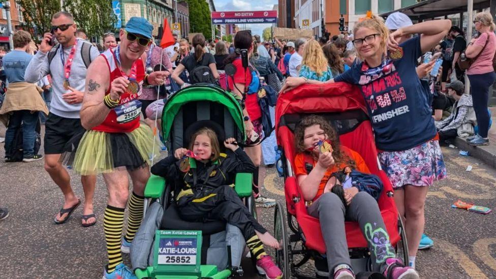A man in a red vest, yellow and black tutu and striped socks, a hat and sunglasses, posing beside a young girl with brown hair and a black top - in a specialist green run chair. They are pictured beside a woman with long blonde hair and glasses, in a navy top and patterned skirt, who is also beside a young brown haired girl in a specialist red run chair. There are numerous marathon goers in the background.