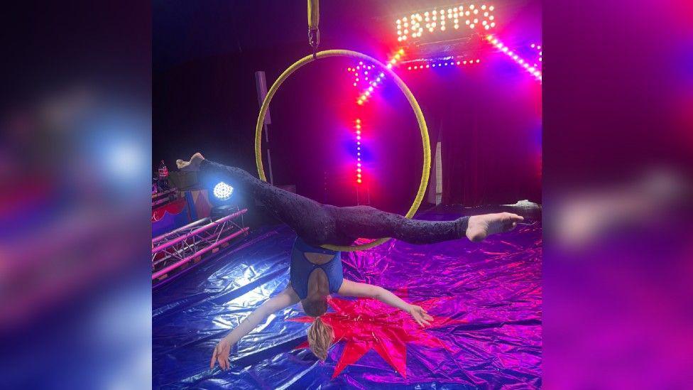 A female acrobat doing the splits upside down hanging from a suspended hoop. The Big Top is dark with purple lighting and the word Devito is lit up in yellow behind the performer.