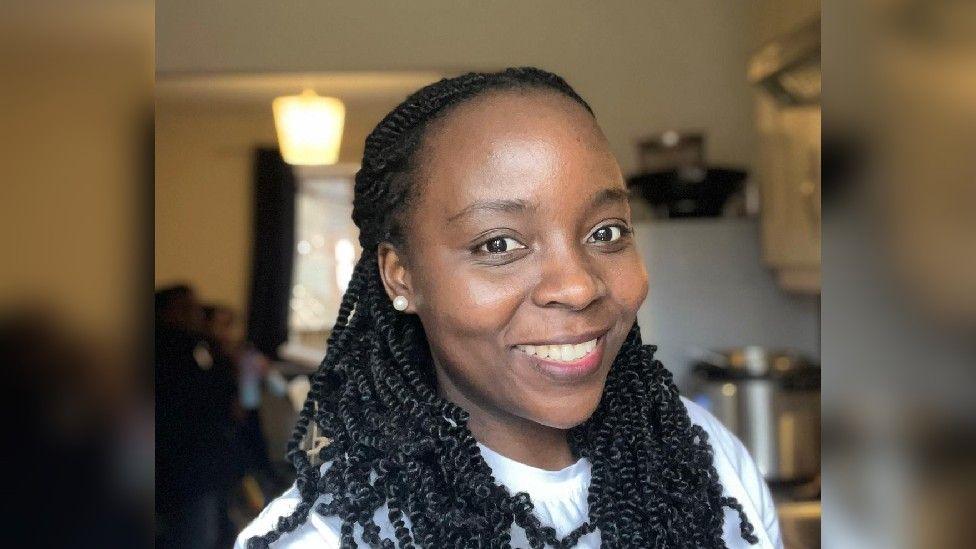 A woman smiles at the camera as she stands inside her home. She is wearing a white top and pearl earrings.