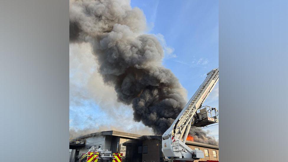 Plumes of black smoke billow into a blue sky as a fire engine can be seen tackling the blaze