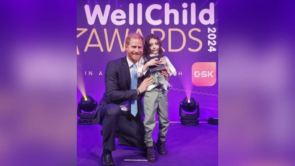 Prince Harry in a suit on a purple stage kneeling down and smiling next to Noah Nicholson who is wearing a grey suit and clutching his award