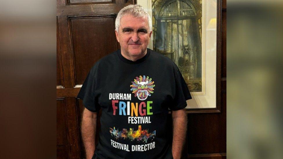 Stephen Cronin standing in front of a picture, wearing a black Durham Fringe T-shirt. He has short grey hair.